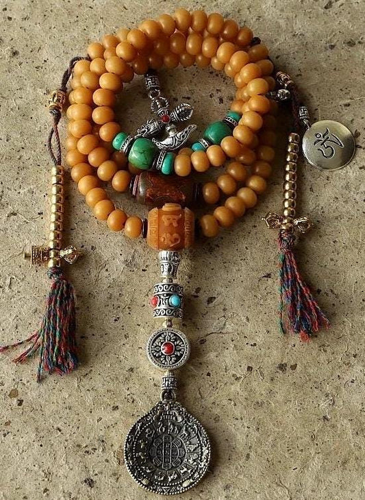 May include: A mala necklace with brown beads, turquoise beads, and silver accents. The necklace has a silver charm with a symbol on it, and a silver tassel. The necklace is on a brown surface.